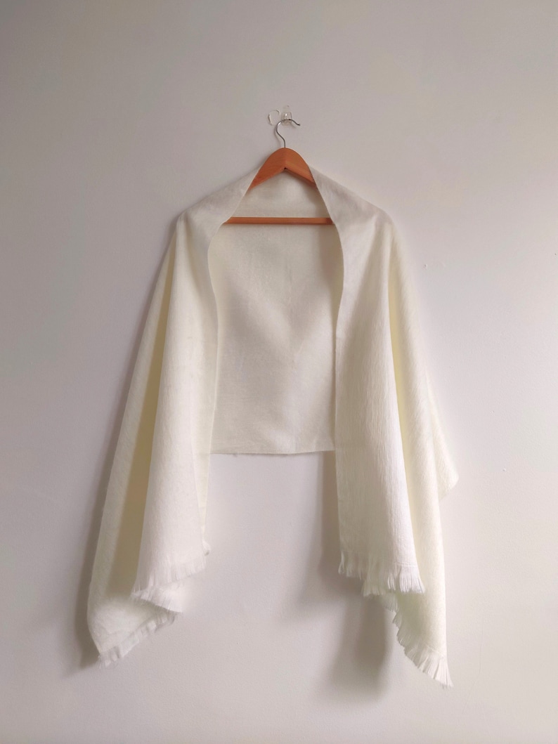 Puede incluir: Un chal rectangular de color blanco roto liso, hecho de una tela ligera y texturizada. La prenda presenta bordes rectos y detalles de flecos cortos y delicados a lo largo del dobladillo inferior, colgado en una percha de madera.