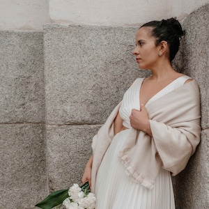 Puede incluir: Una persona con un vestido y chal crema, sosteniendo un ramo de flores blancas. El vestido tiene una falda plisada y un escote en V. El chal está drapeado sobre los hombros. El fondo es una pared de piedra.