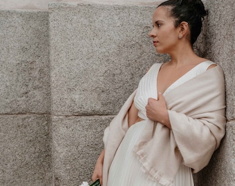 May include: A woman in a cream-colored dress and shawl, holding a bouquet of white flowers. The dress has a pleated skirt and a V-neckline. The shawl is draped over the shoulders. The background is a stone wall.