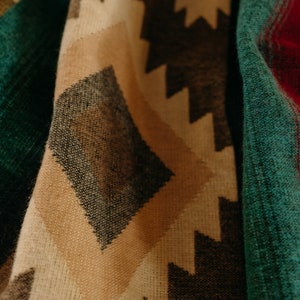 Op de afbeelding: Close-up van een gevouwen textiel met een geometrisch patroon. Het ontwerp heeft ruit- en zigzagvormen in tinten bruin, bruin, blauwgroen, rood en goud. De stof heeft een geweven textuur, wat wijst op een deken of sjaal.