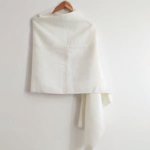 Puede incluir: Un chal rectangular de color blanco roto liso drapeado sobre una percha de madera. La tela tiene una textura suave y mate, y cuelga con un pliegue suave en un lado, creando una apariencia limpia y sencilla sobre un fondo claro y liso.