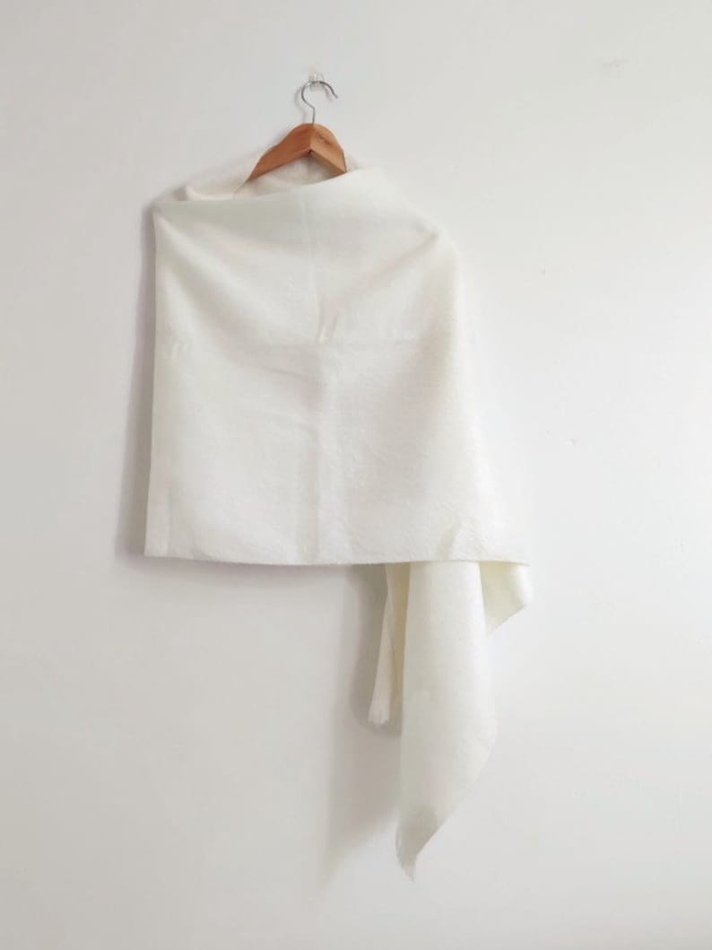 Puede incluir: Un chal rectangular de color blanco roto liso drapeado sobre una percha de madera. La tela tiene una textura suave y mate, y cuelga con un pliegue suave en un lado, creando una apariencia limpia y sencilla sobre un fondo claro y liso.