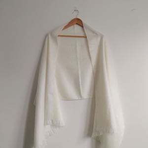 Puede incluir: Un chal rectangular de color blanco roto liso con una apariencia suave y texturizada, y detalles de flecos delicados en los bordes inferiores. La prenda cuelga de una percha de madera, mostrando su forma larga, fluida y tejido ligero.