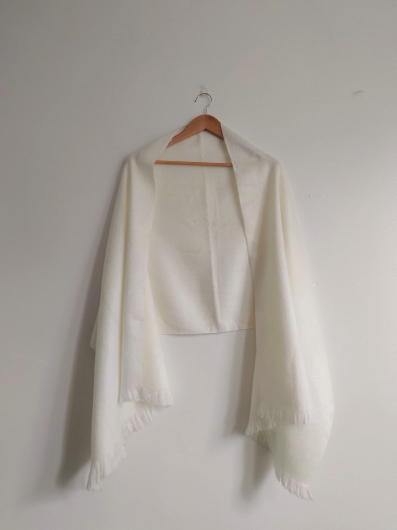 Puede incluir: Un chal rectangular de color blanco roto liso con una apariencia suave y texturizada, y detalles de flecos delicados en los bordes inferiores. La prenda cuelga de una percha de madera, mostrando su forma larga, fluida y tejido ligero.