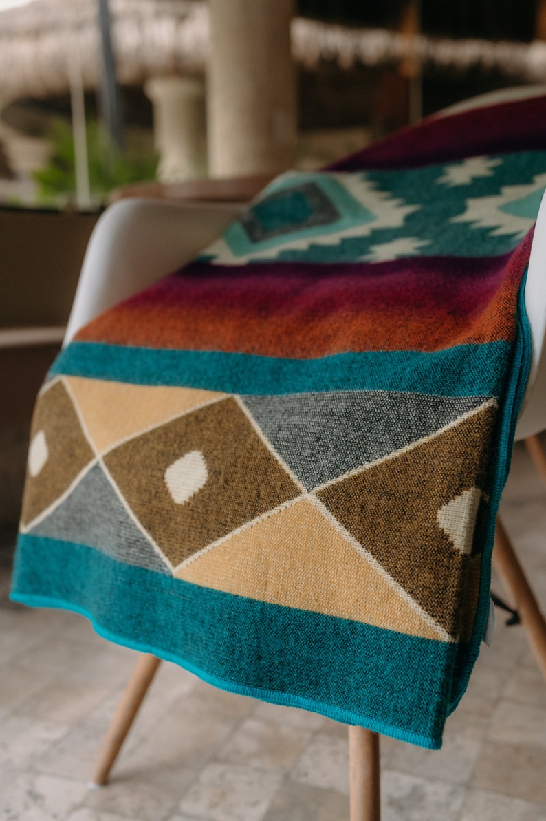 K&ouml;nnte beinhalten: Eine gewebte Wohndecke mit einem geometrischen Muster aus Rauten und Dreiecken in den Farben Petrol, Senfgelb, Dunkelbraun und tiefem Magenta. Das Textil hat eine weiche Struktur und einen ges&auml;umten Rand und zeigt eine lebendige Farbpalette.