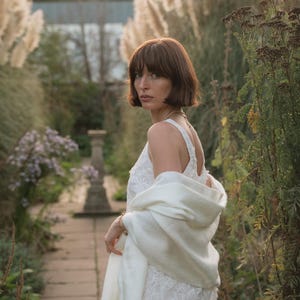 Puede incluir: Un vestido blanco sin mangas con un intrincado encaje floral superpuesto y un chal de punto blanco a juego drapeado sobre los hombros. El vestido tiene un escote redondo y un dobladillo recto, mientras que el chal presenta una textura suave.