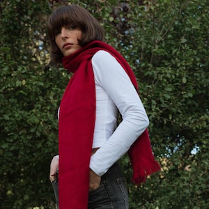 Puede incluir: Una bufanda larga de color rojo sólido con extremos deshilachados, usada sobre una camisa blanca de manga larga y pantalones vaqueros de color gris oscuro. La bufanda está envuelta alrededor del cuello y cae al frente con textura tejida.