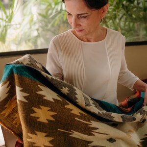 K&ouml;nnte beinhalten: Eine gewebte Decke mit einem geometrischen Muster in Braun-, Creme-, Petrol- und Gelbt&ouml;nen. Das Textil zeigt sich wiederholende sternartige Motive und horizontale Streifen, die eine weiche Textur und ein mehrfarbiges Design auf der Oberfl&auml;che aufweisen.