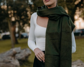 May include: A woman wearing a white long-sleeved shirt and a dark green scarf. The scarf is wrapped around her neck and tied in a loose knot. She is standing in a park with trees and grass in the background.