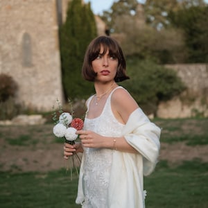 Puede incluir: Un vestido blanco sin mangas con intrincados bordados florales y escote cuadrado, combinado con un suave chal color crema con flecos. El conjunto se complementa con un delicado collar dorado y un peque&ntilde;o ramo de flores blancas y rojas.