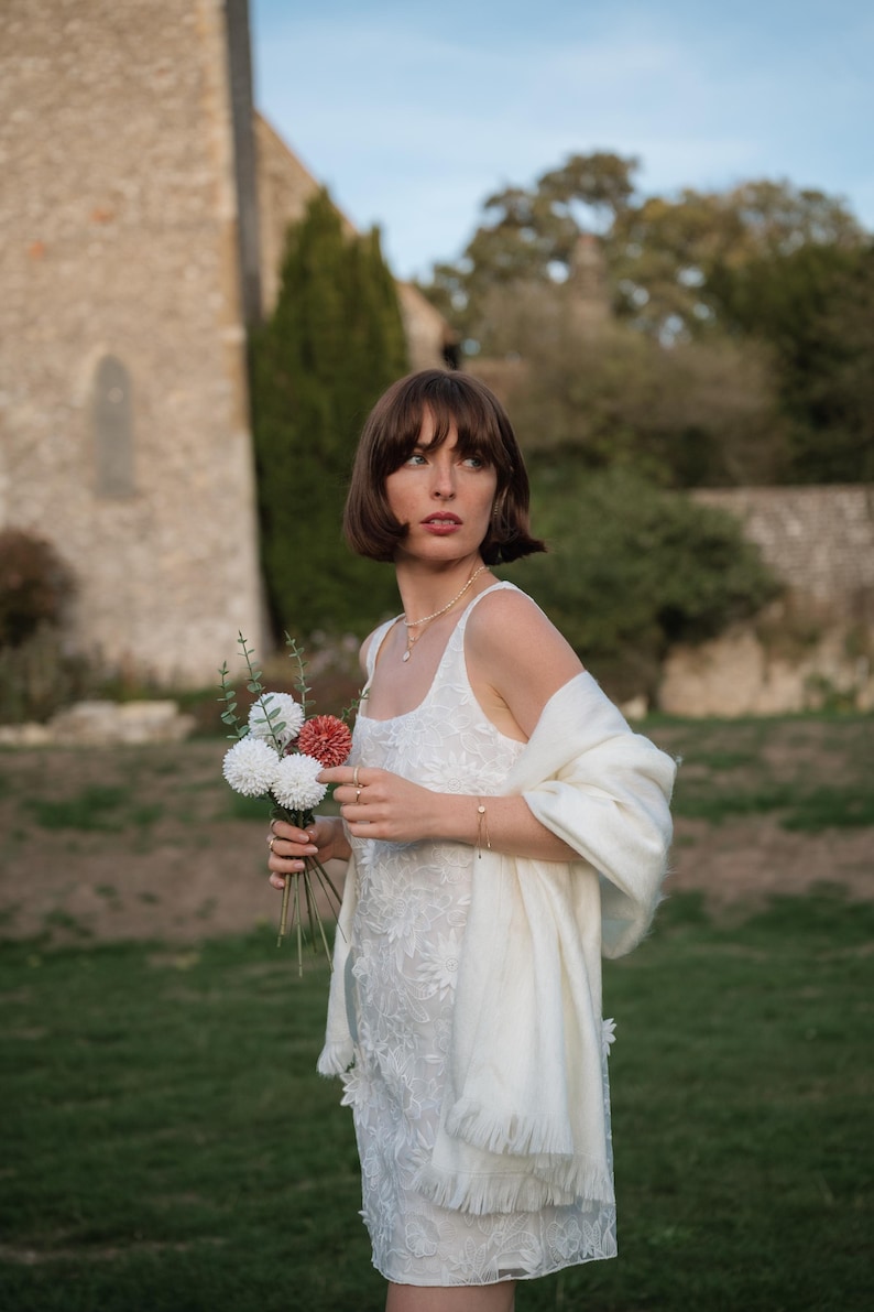Puede incluir: Un vestido blanco sin mangas con intrincados bordados florales y escote cuadrado, combinado con un suave chal color crema con flecos. El conjunto se complementa con un delicado collar dorado y un peque&ntilde;o ramo de flores blancas y rojas.