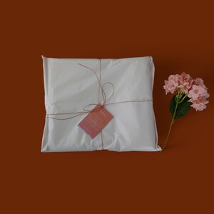 K&ouml;nnte beinhalten: Eine wei&szlig;e Geschenkt&uuml;te, die mit Schnur gebunden ist und einen kleinen Dankesanh&auml;nger hat. Die T&uuml;te liegt auf einem braunen Hintergrund mit einer rosa Blume.