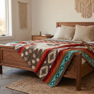 Op de afbeelding: Een houten bedframe met een deken met geometrische patronen in tinten rood, oranje, bruin, wit en turquoise. De deken heeft een centraal ruitmotief en gestreepte randen, gedrapeerd over wit beddengoed met beige kussens.