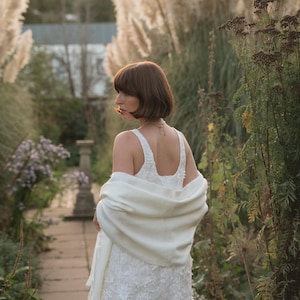 Puede incluir: Un vestido blanco sin mangas con intrincados bordados florales y escote redondo, combinado con un chal blanco liso y suave sobre los hombros. La prenda presenta una apariencia texturizada con detallados patrones bot&aacute;nicos.