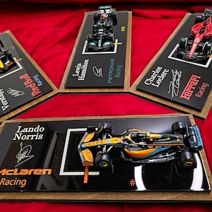 Puede incluir: Cuatro modelos de coches de carreras de F&oacute;rmula 1 montados sobre placas de madera rectangulares con bases de acr&iacute;lico negro. Cada expositor muestra el nombre del piloto, el equipo, el n&uacute;mero de carrera y una firma impresa.