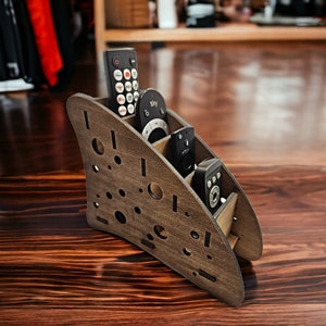 May include: A wooden remote control organiser with a unique design. It has four slots for remotes and a cutout in the shape of a wave. The organiser is made of dark wood and has a natural finish.