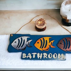 Puede incluir: Un letrero de madera azul con la palabra "BATHROOM" escrita en pintura blanca. El letrero tiene tres recortes de peces coloridos: un pez plateado, un pez naranja y un pez marrón.