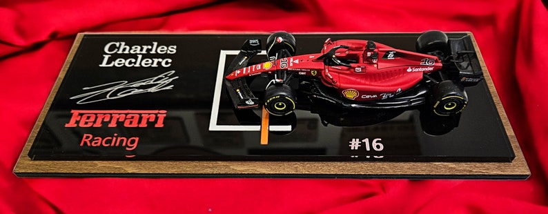 Puede incluir: Un coche de carreras modelo Ferrari de F&oacute;rmula 1 rojo montado sobre una placa de exhibici&oacute;n negra. La placa presenta el texto Charles Leclerc, una firma, el logotipo de Ferrari Racing y el n&uacute;mero 16. El coche modelo tiene neum&aacute;ticos negros.