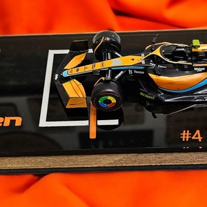 Puede incluir: Un modelo a escala de un coche de carreras de F&oacute;rmula 1 McLaren, n&uacute;mero 4, conducido por Lando Norris. El modelo est&aacute; montado sobre una base de exhibici&oacute;n rectangular negra con el texto Lando Norris, McLaren Racing y una firma impresa.