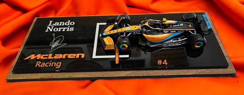 Puede incluir: Un modelo a escala de un coche de carreras de F&oacute;rmula 1 McLaren, n&uacute;mero 4, conducido por Lando Norris. El modelo est&aacute; montado sobre una base de exhibici&oacute;n rectangular negra con el texto Lando Norris, McLaren Racing y una firma impresa.