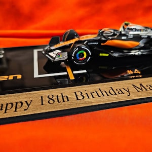 Puede incluir: Un modelo a escala de coche de carreras McLaren de F&oacute;rmula 1 con la marca Lando Norris, expuesto sobre una base negra con el texto Lando Norris, McLaren Racing, #4, y una placa de madera grabada Happy 18th Birthday Max.