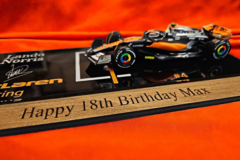 Puede incluir: Un modelo a escala de coche de carreras McLaren de F&oacute;rmula 1 con la marca Lando Norris, expuesto sobre una base negra con el texto Lando Norris, McLaren Racing, #4, y una placa de madera grabada Happy 18th Birthday Max.