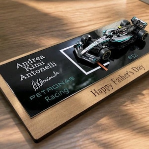 Puede incluir: Un modelo de coche de carreras de F&oacute;rmula 1 en miniatura montado sobre una base rectangular de madera. La placa negra muestra el texto Andrea Kimi Antonelli, una firma, Petronas Racing y el n&uacute;mero 12. La base de madera tiene grabado Happy Father's Day.