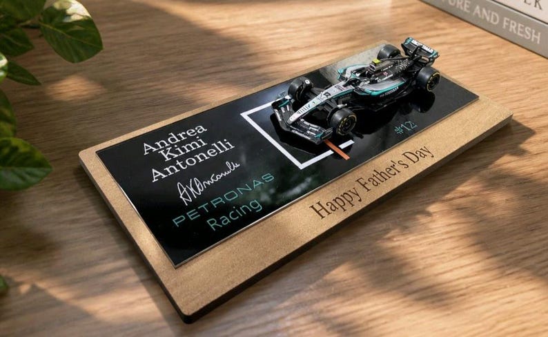 Puede incluir: Un modelo de coche de carreras de F&oacute;rmula 1 en miniatura montado sobre una base rectangular de madera. La placa negra muestra el texto Andrea Kimi Antonelli, una firma, Petronas Racing y el n&uacute;mero 12. La base de madera tiene grabado Happy Father's Day.