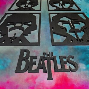 May include: Black wooden wall art featuring four square panels with band member silhouettes and the band name "The Beatles". The art is set against a colorful, abstract background.