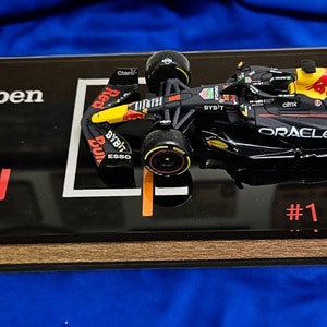 Puede incluir: Un modelo a escala de un coche de F&oacute;rmula 1 de Red Bull Racing en negro y azul oscuro con el n&uacute;mero 1, marcas de Oracle y Bybit, y una toma de aire amarilla. El modelo est&aacute; montado sobre una placa negra con el texto Max Verstappen, Red Bull Racing y una firma.