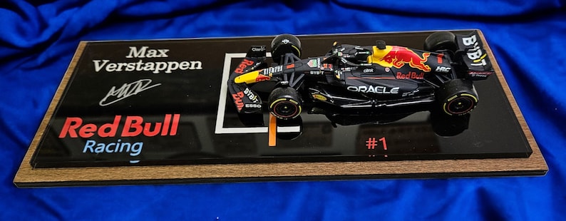 Puede incluir: Un modelo a escala de un coche de F&oacute;rmula 1 de Red Bull Racing en negro y azul oscuro con el n&uacute;mero 1, marcas de Oracle y Bybit, y una toma de aire amarilla. El modelo est&aacute; montado sobre una placa negra con el texto Max Verstappen, Red Bull Racing y una firma.