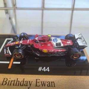 Puede incluir: Un modelo de coche de carreras de F&oacute;rmula 1 rojo y blanco con el n&uacute;mero 44 y logotipos de HP y Shell. El coche est&aacute; montado sobre una base negra con el texto #44 y Birthday Ewan impreso en el borde frontal, mostrando neum&aacute;ticos negros.