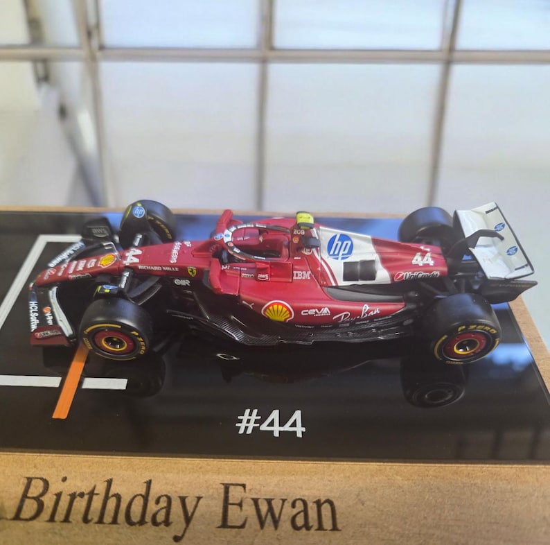 Puede incluir: Un modelo de coche de carreras de F&oacute;rmula 1 rojo y blanco con el n&uacute;mero 44 y logotipos de HP y Shell. El coche est&aacute; montado sobre una base negra con el texto #44 y Birthday Ewan impreso en el borde frontal, mostrando neum&aacute;ticos negros.