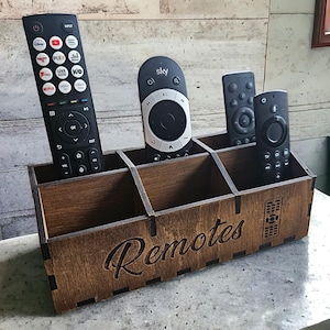 May include: A wooden remote control organiser with the word "Remotes" engraved on the front. The organiser has six slots for remotes and is made of dark brown wood.