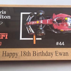 Puede incluir: Una placa de exhibici&oacute;n rectangular de madera con un modelo de coche de carreras Ferrari de F&oacute;rmula 1 rojo con el n&uacute;mero 44. La superficie negra incluye el texto Lewis Hamilton, Ferrari Racing, #44, una firma y el mensaje Happy 18th Birthday Ewan.