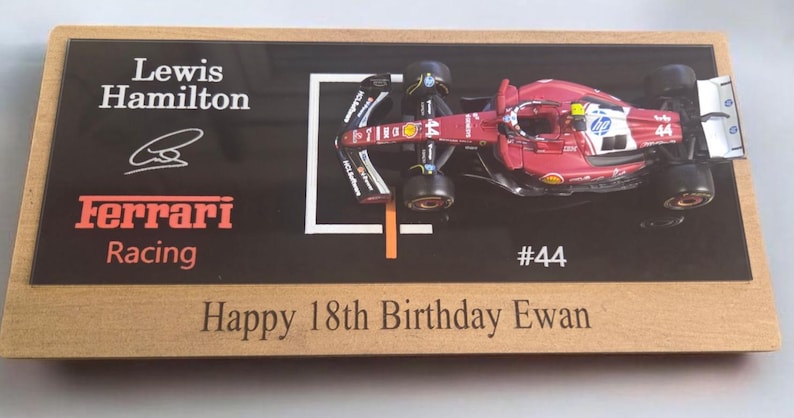 Puede incluir: Una placa de exhibici&oacute;n rectangular de madera con un modelo de coche de carreras Ferrari de F&oacute;rmula 1 rojo con el n&uacute;mero 44. La superficie negra incluye el texto Lewis Hamilton, Ferrari Racing, #44, una firma y el mensaje Happy 18th Birthday Ewan.