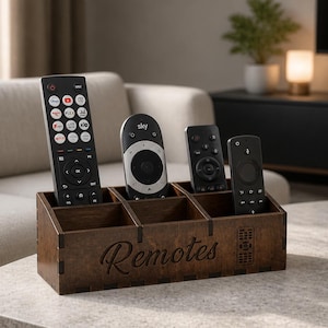 Puede incluir: Un organizador de control remoto de madera con cuatro compartimentos, que contiene cuatro controles remotos negros. El organizador es marrón con la palabra "Remotes" grabada en la parte delantera. Los controles remotos son de varios tamaños y diseños.