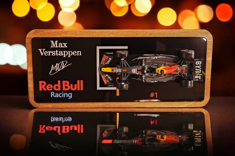 Puede incluir: Una placa de exhibici&oacute;n rectangular de madera con un modelo en miniatura de un coche de F&oacute;rmula 1 de Red Bull Racing en azul oscuro y rojo. La superficie negra incluye el texto Max Verstappen, una firma, Red Bull Racing y el n&uacute;mero 1.