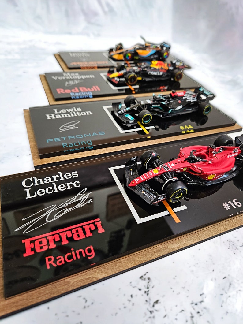 Puede incluir: Una colecci&oacute;n de cuatro coches de carreras de F&oacute;rmula 1 a escala montados sobre placas de exhibici&oacute;n negras individuales con bases de madera. Cada placa muestra el nombre del piloto, una firma y el logotipo del equipo, incluyendo Ferrari, Mercedes, Red Bull y McLaren.