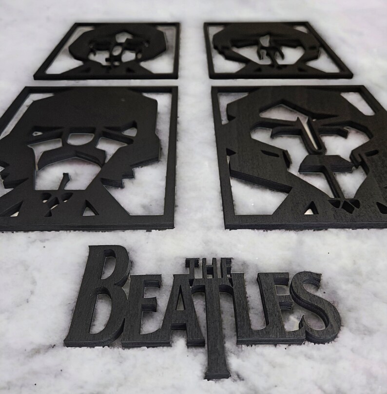 May include: Black wooden coasters featuring cut-out portraits of The Beatles band members. The word "THE BEATLES" is also displayed in a matching font. The coasters are square and the text is in a serif font.