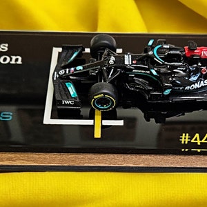 Puede incluir: Un modelo en miniatura de coche de carreras de F&oacute;rmula 1 Mercedes-AMG Petronas negro montado sobre una placa de exhibici&oacute;n rectangular de acr&iacute;lico negro con base de madera. La placa presenta el texto Lewis Hamilton, Petronas Racing y #44 con una firma.