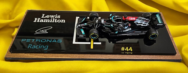 Puede incluir: Un modelo en miniatura de coche de carreras de F&oacute;rmula 1 Mercedes-AMG Petronas negro montado sobre una placa de exhibici&oacute;n rectangular de acr&iacute;lico negro con base de madera. La placa presenta el texto Lewis Hamilton, Petronas Racing y #44 con una firma.