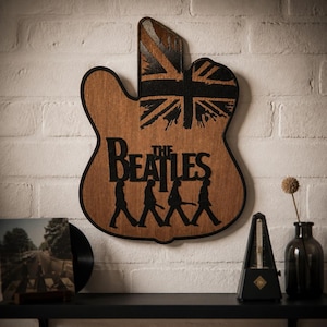 Puede incluir: Un adorno de pared de madera con forma de cuerpo de guitarra que presenta un diseño de Union Jack negro en la parte superior y el texto negro The Beatles sobre una silueta de los cuatro miembros de la banda caminando en fila, bordeado en negro.