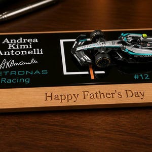 Puede incluir: Una placa de exhibici&oacute;n rectangular de madera con un modelo en miniatura de coche de carreras de F&oacute;rmula 1 en negro y plata. La pieza de acr&iacute;lico negro muestra el texto Andrea Kimi Antonelli, una firma, Petronas Racing y #12. La base de madera tiene grabado Happy Father's Day.