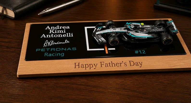 Puede incluir: Una placa de exhibici&oacute;n rectangular de madera con un modelo en miniatura de coche de carreras de F&oacute;rmula 1 en negro y plata. La pieza de acr&iacute;lico negro muestra el texto Andrea Kimi Antonelli, una firma, Petronas Racing y #12. La base de madera tiene grabado Happy Father's Day.