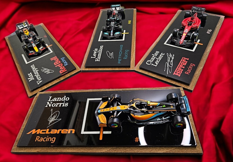 Puede incluir: Cuatro modelos de coches de carreras de F&oacute;rmula 1 montados sobre placas de madera rectangulares con bases de acr&iacute;lico negro. Cada expositor muestra el nombre del piloto, el equipo, el n&uacute;mero de carrera y una firma impresa.