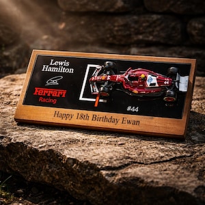 Puede incluir: Una placa rectangular de madera con un modelo a escala de un coche de carreras Ferrari de F&oacute;rmula 1 rojo. La superficie negra incluye el texto Lewis Hamilton, Ferrari Racing, #44 y la inscripci&oacute;n grabada Happy 18th Birthday Ewan.
