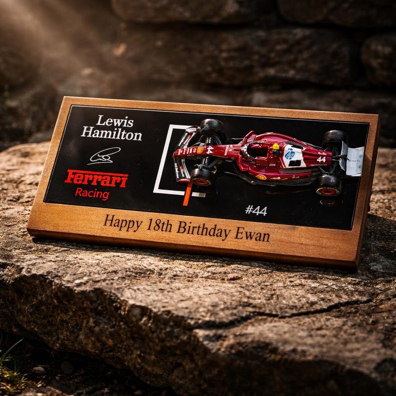 Puede incluir: Una placa rectangular de madera con un modelo a escala de un coche de carreras Ferrari de F&oacute;rmula 1 rojo. La superficie negra incluye el texto Lewis Hamilton, Ferrari Racing, #44 y la inscripci&oacute;n grabada Happy 18th Birthday Ewan.