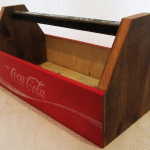 Könnte beinhalten: Eine Holzkiste mit einem roten Coca-Cola-Logo, das auf die Vorderseite gemalt ist. Die Holzkiste hat einen schwarzen Griff und ist aus Naturholz gefertigt.