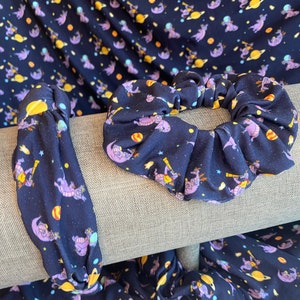 May include: Two hair scrunchies with a dark blue background and a repeating pattern of purple dragons, stars, and planets. The scrunchies are made of a soft, stretchy fabric.